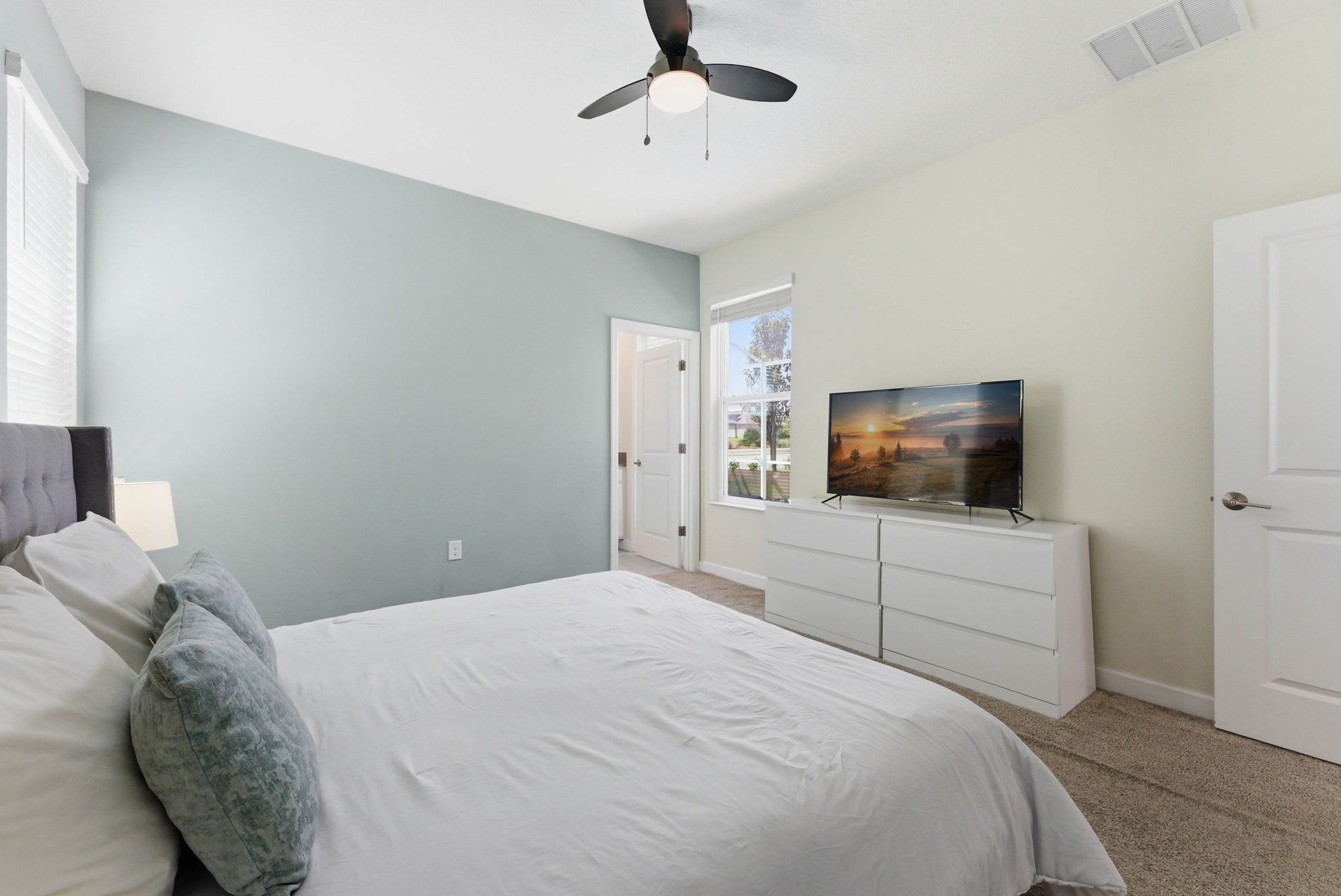 Une chambre à coucher avec un lit, un téléviseur posé sur un meuble, un ventilateur de plafond et une porte donnant sur une autre pièce.