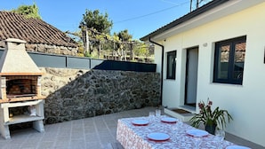 Uma área de refeições ao ar livre com uma churrasqueira de pedra, uma mesa posta com pratos e copos, e uma casa com telhado de cerâmica ao fundo.