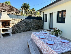 An outdoor dining area with a stone barbecue grill, a table set with plates and glasses, and a house with a tiled roof in the background. - Holiday Home 'Quinta De Fonte Coberta' with Mountain View, Wi-Fi and Air Conditioning (Lavradas)