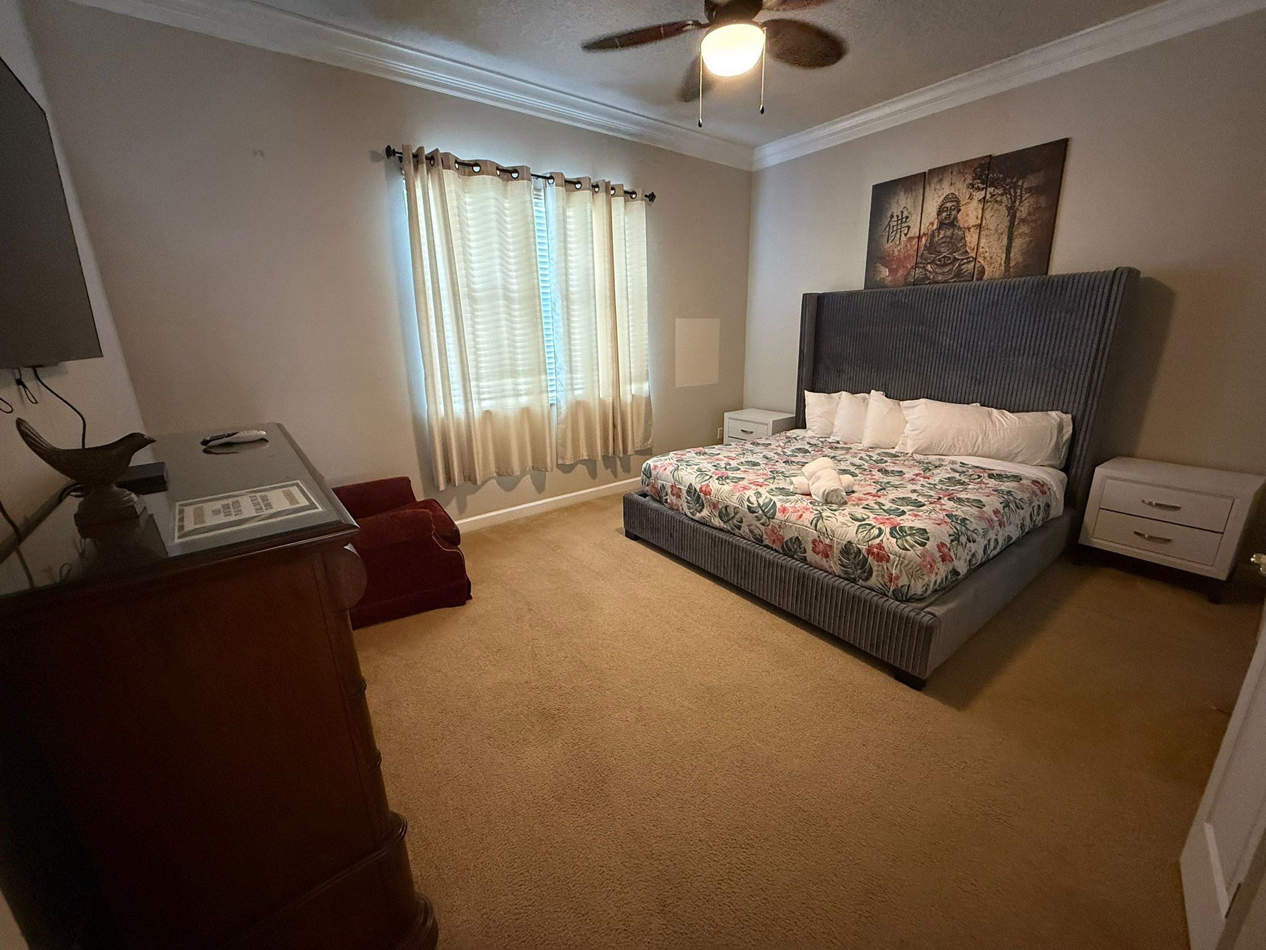 Un dormitorio con cama, mesitas de noche, escritorio, silla, ventilador de techo y ventana con cortinas.