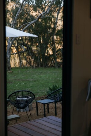 Eine Terrasse im Freien mit einem Holztisch und zwei Stühlen, einem weißen Sonnenschirm und Blick auf eine grüne Fläche mit Bäumen.