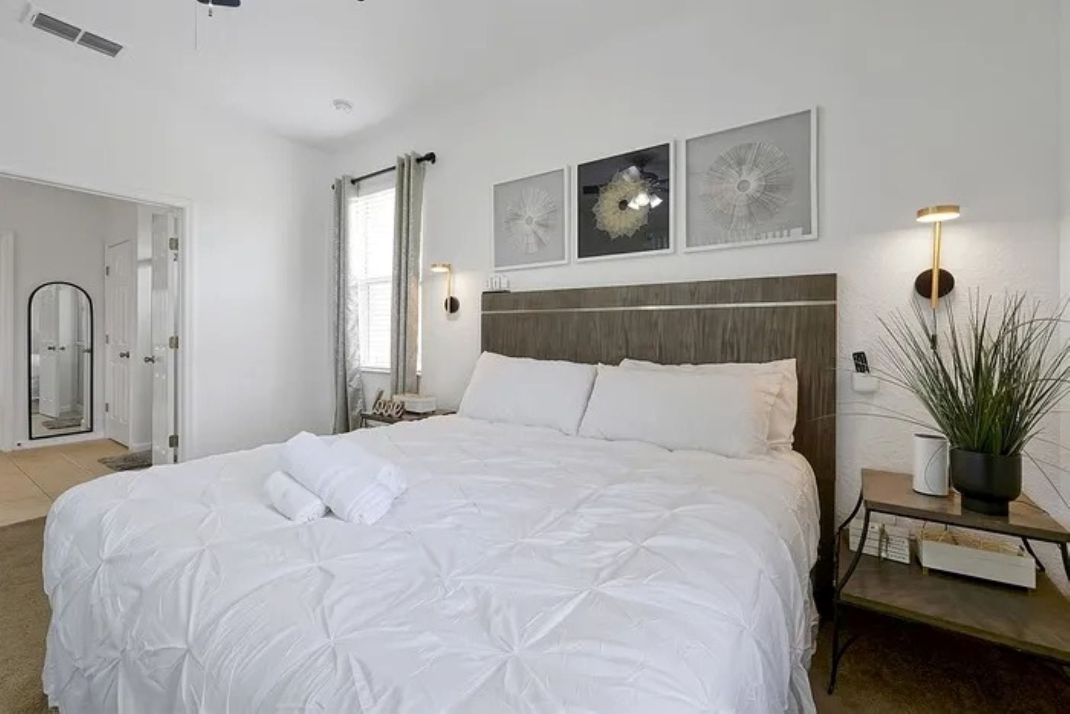 En pænt redt seng med hvide sengetæpper, et natbord med lampe og en potteplante.