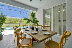 Taula de dinar de fusta amb cadires, una planta en test i vetreria a un balcó amb vista a una piscina i vegetació.
