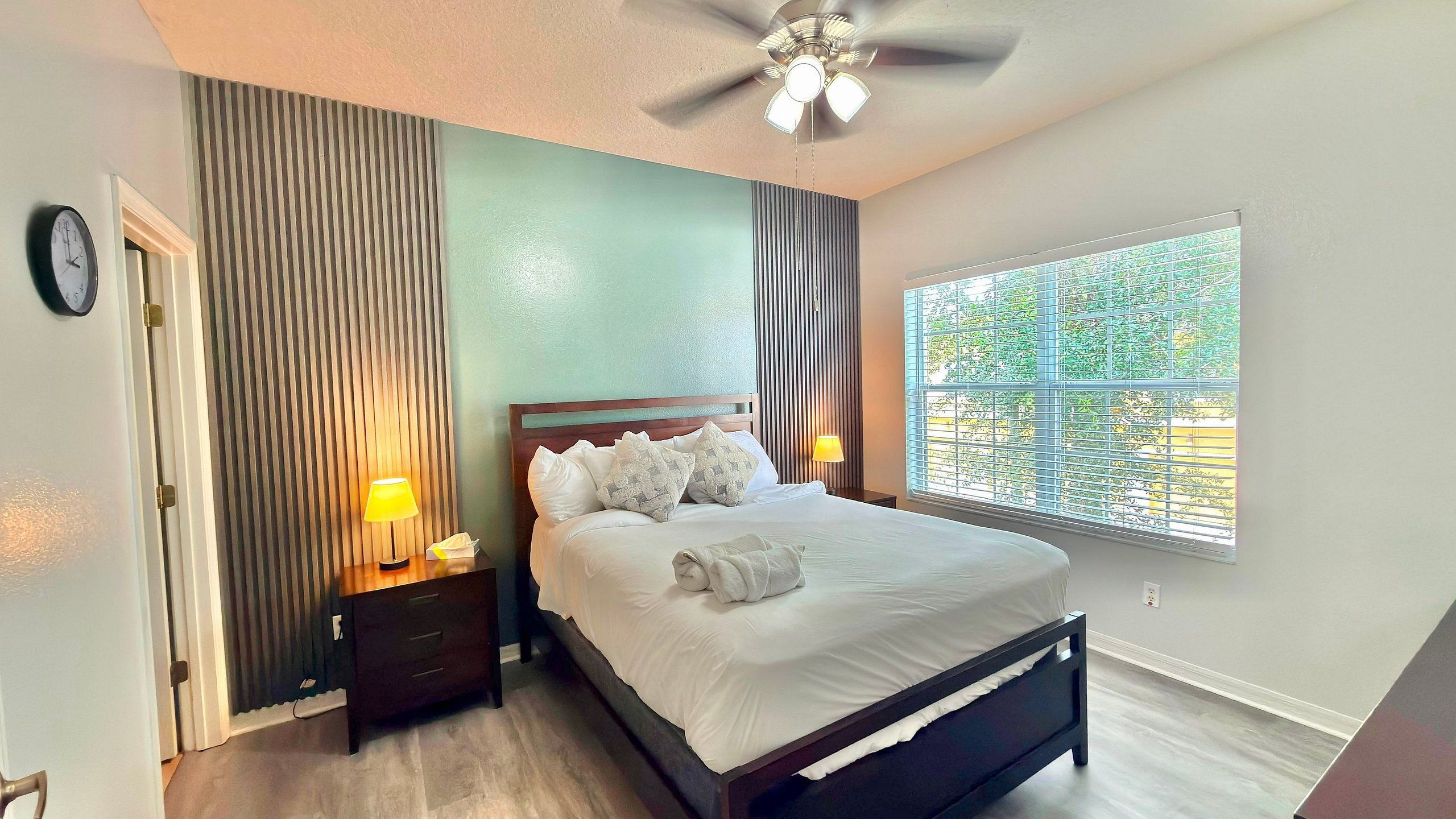 A bedroom with a bed, nightstands, a lamp, a ceiling fan, a window with blinds, and a clock on the wall.