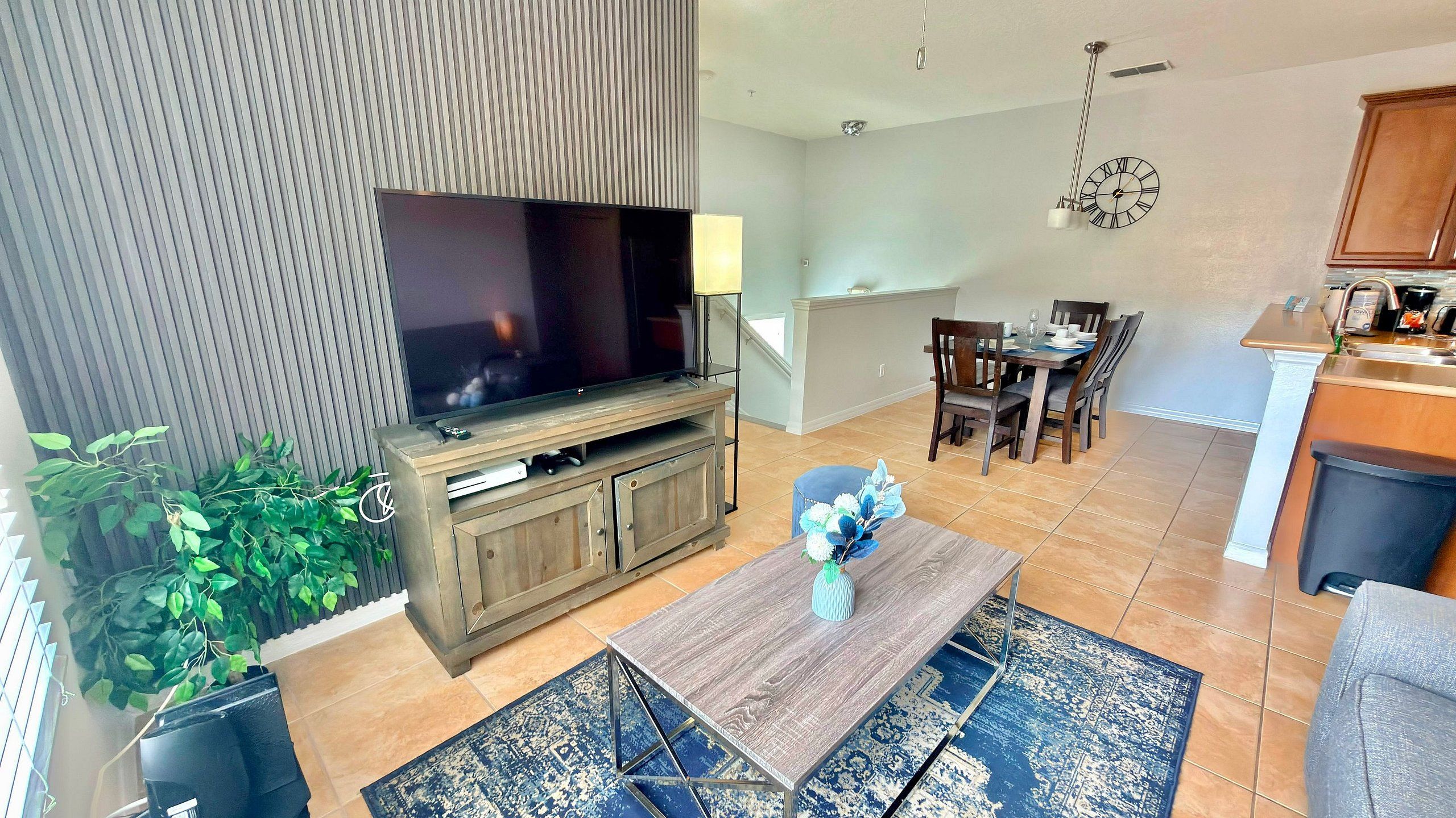 Una sala moderna con televisor de pantalla plana, mueble de entretenimiento de madera, comedor con sillas y mesa, cocina con mostrador y fregadero, y una alfombra azul estampada.