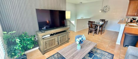 Una sala moderna con televisor de pantalla plana, mueble de entretenimiento de madera, comedor con sillas y mesa, cocina con mostrador y fregadero, y una alfombra azul estampada.