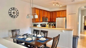 Un comedor con una mesa dispuesta para cuatro, una cocina con armarios de madera y un reloj de pared.