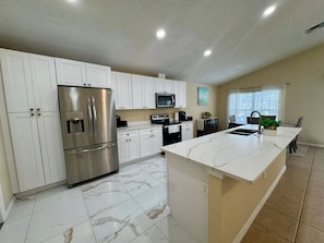 A modern kitchen with white cabinetry, a stainless steel refrigerator, a microwave, and a marble island. - Lake Berkley 3BR – Disney 10min, Pool & Lake (Kissimmee)