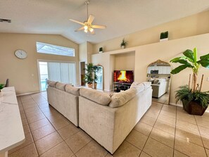 A living room with a beige sofa, a flat-screen TV, a ceiling fan, and a kitchen area visible through an open door. - Lake Berkley 3BR – Disney 10min, Pool & Lake (Kissimmee)