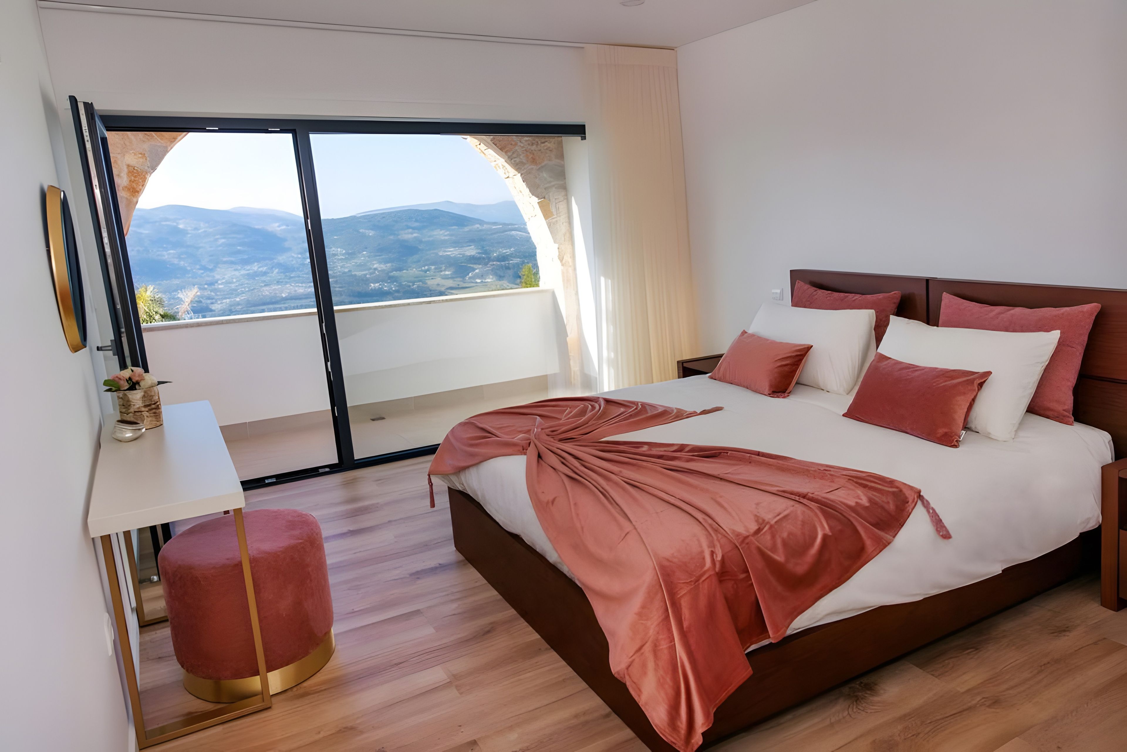 A modern bedroom with a large bed, a bedside table, a lamp, and a view of the mountains through a glass door.