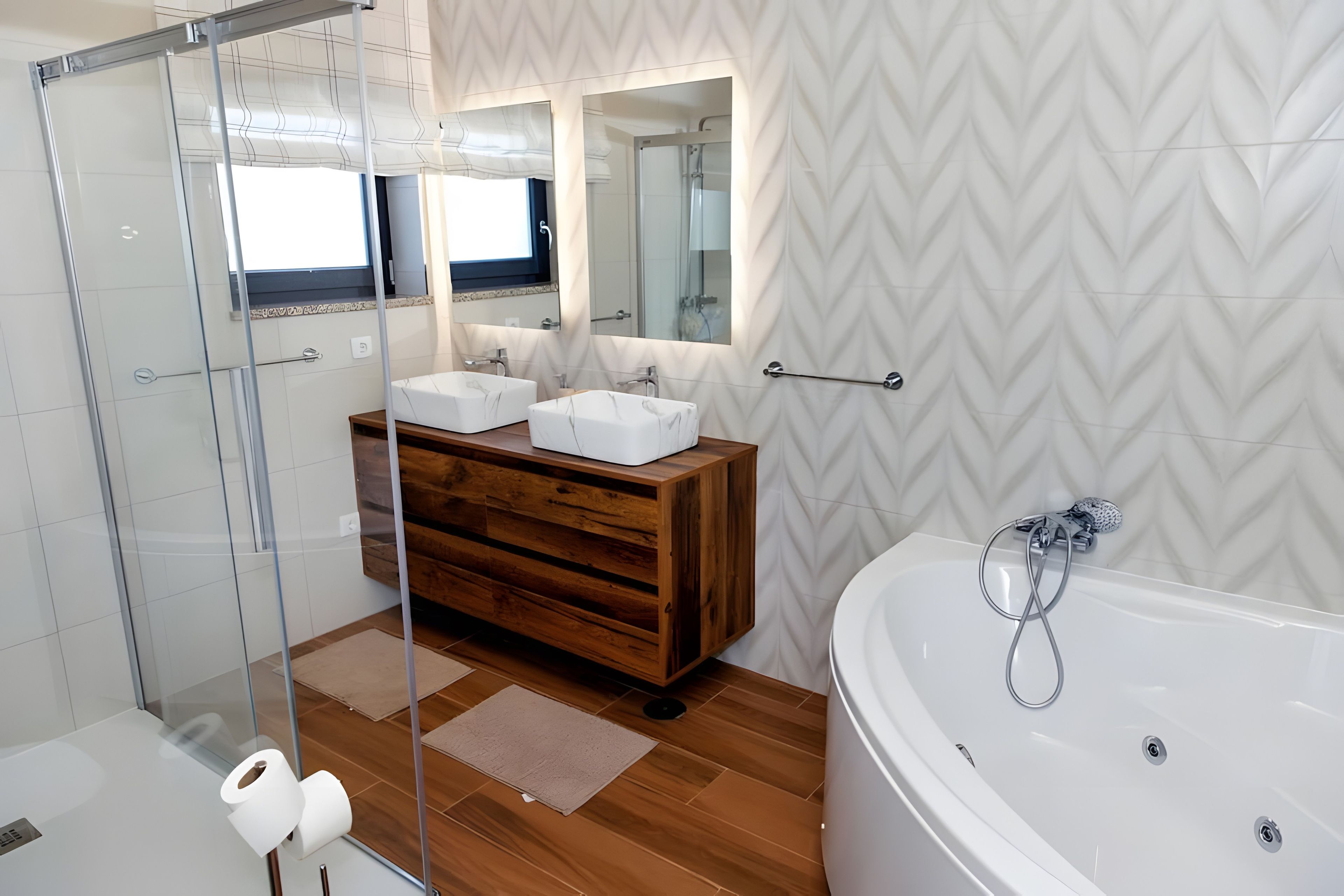 Une salle de bain moderne dotée d’une douche à parois de verre, d’un meuble-lavabo en bois avec deux vasques, d’une grande baignoire et d’un mur orné d’un motif en chevrons.
