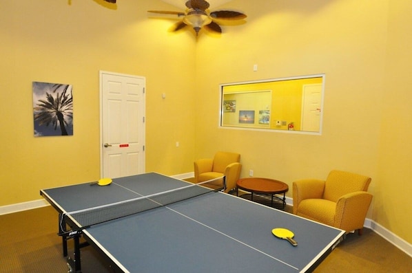 Ein Zimmer mit Tischtennisplatte, zwei gelben Sesseln, einem Deckenventilator und einer Tür.