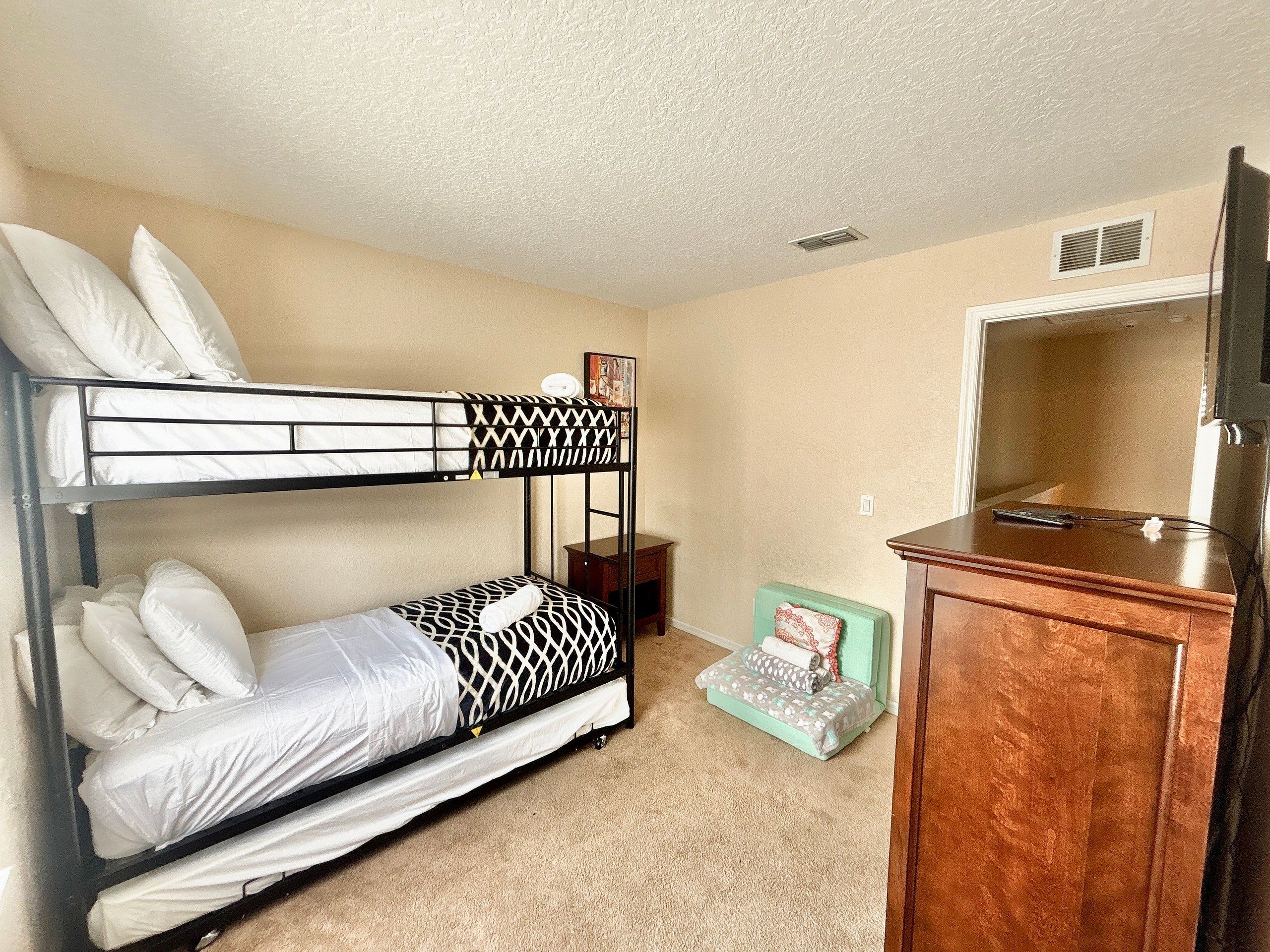 A bunk bed with white bedding, a small wooden nightstand, a green cushioned bench, and a wooden cabinet with a television.