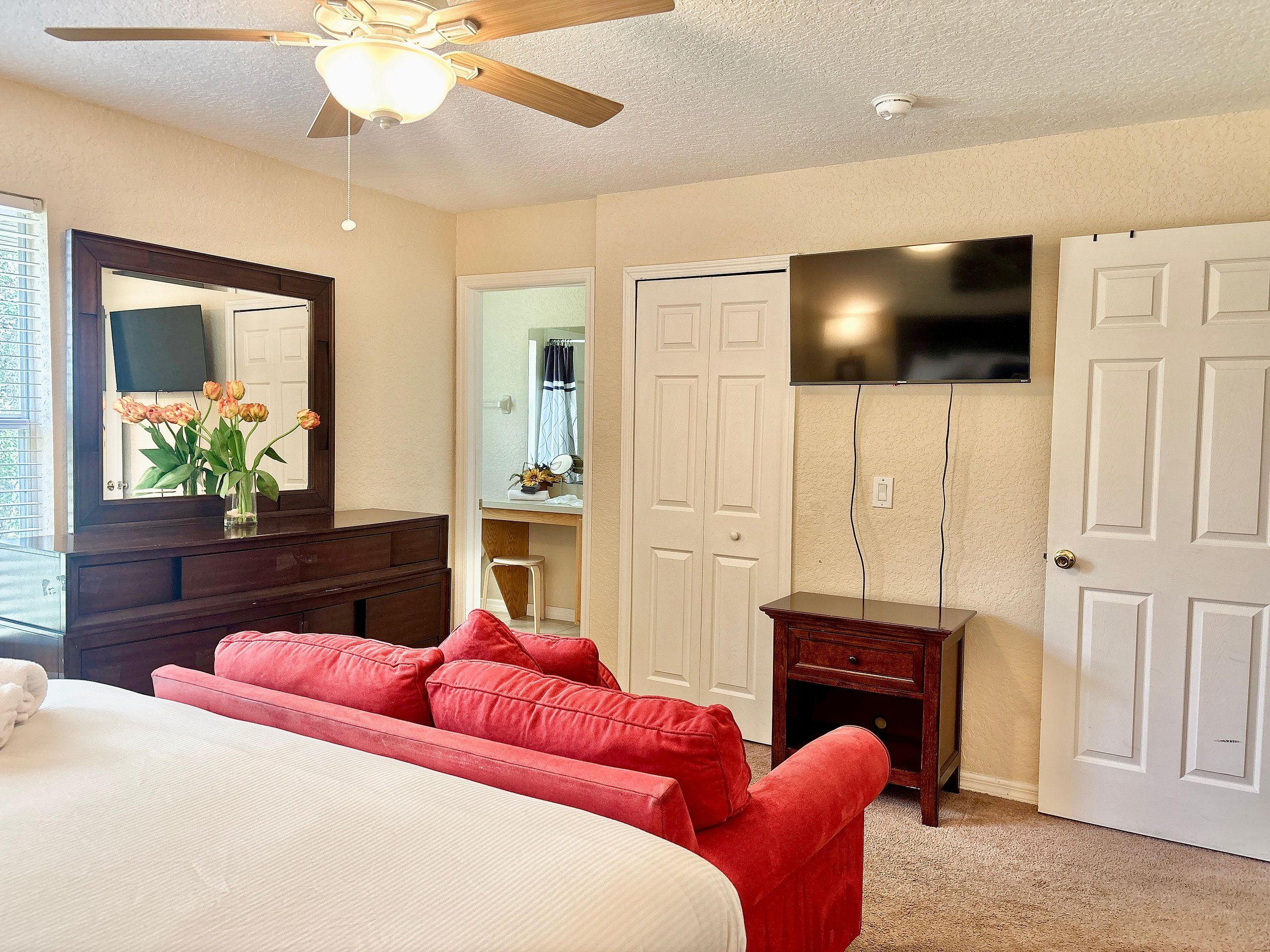 A bedroom with a bed, a red sofa, a ceiling fan, a TV mounted on the wall, and a door.