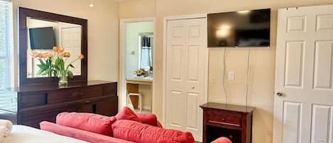 A bedroom with a bed, a red sofa, a ceiling fan, a TV mounted on the wall, and a door.