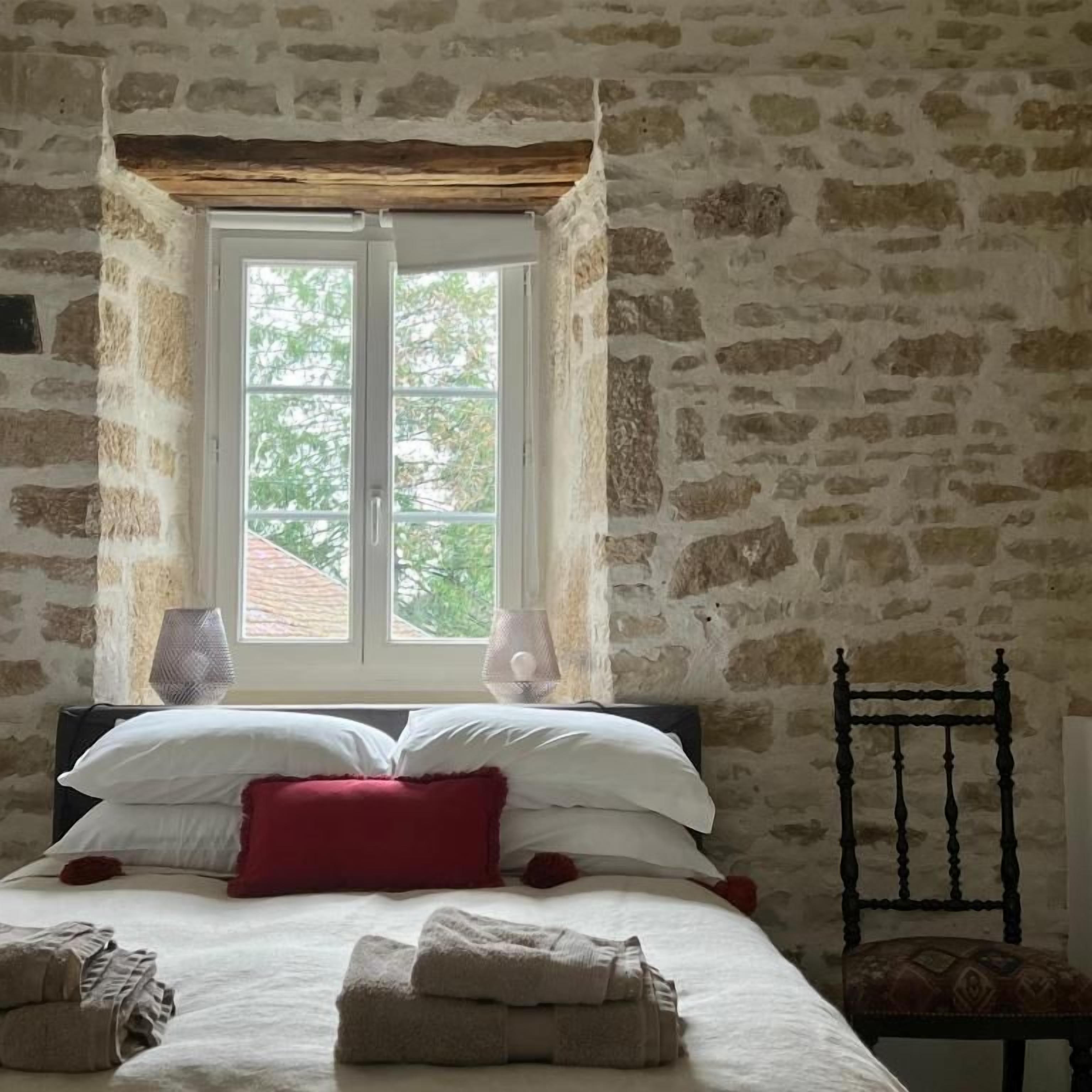 A room with a stone wall, a bed with white and red bedding, a chair, and a window with a view of greenery.