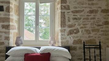 A room with a stone wall, a bed with white and red bedding, a chair, and a window with a view of greenery.