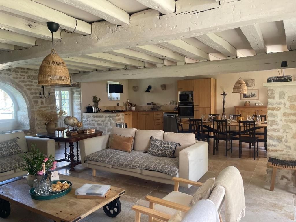 A spacious living area with a sofa, coffee table, and dining table. A kitchen with wooden cabinets and a stone wall is visible in the background.