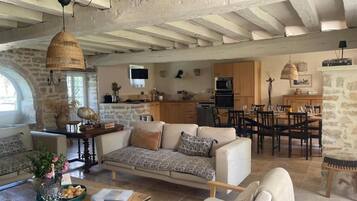 A spacious living area with a sofa, coffee table, and dining table. A kitchen with wooden cabinets and a stone wall is visible in the background.