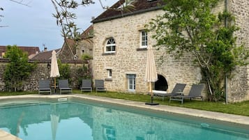A swimming pool with lounge chairs and umbrellas, surrounded by stone buildings with windows and a clear sky.