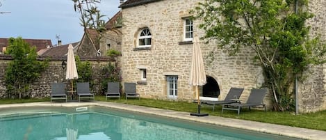 A swimming pool with lounge chairs and umbrellas, surrounded by stone buildings with windows and a clear sky.
