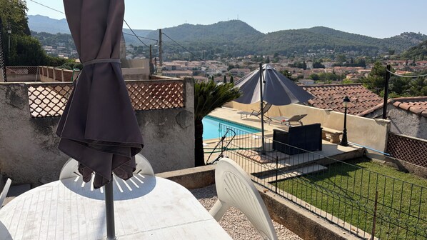 A terrace with a pool, outdoor furniture, and a view of a town and mountains.