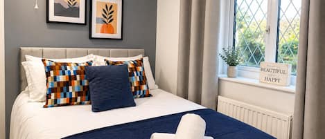 Ein Schlafzimmer mit einem Bett, Kissen, einer Decke, einer Hängeleuchte, einem Fenster mit Vorhängen und zwei gerahmten Bildern an der Wand.
