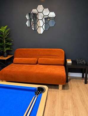 Une pièce moderne équipée d’une table de billard, d’un canapé, d’un miroir hexagonal et d’une petite table d’appoint agrémentée d’une plante.
