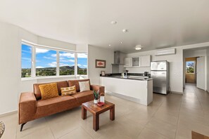 A modern living space with a sofa, coffee table, kitchen area, and large windows offering a view of the outdoors. - Blue Bay Ocean View | Luxury 2-Bedroom with Beach Access (Willemstad)
