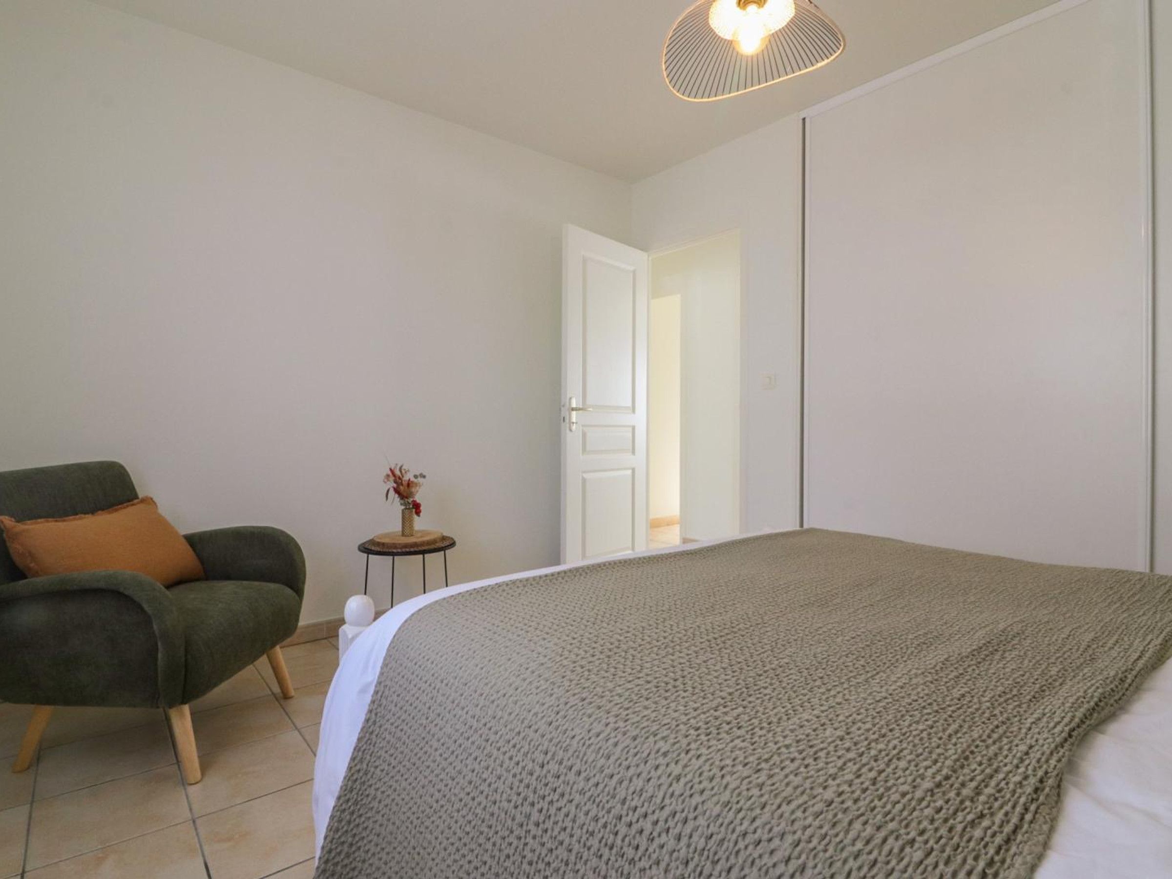 Une chambre à coucher comprenant un lit, une chaise, une petite table avec un vase et une porte donnant sur une autre pièce.