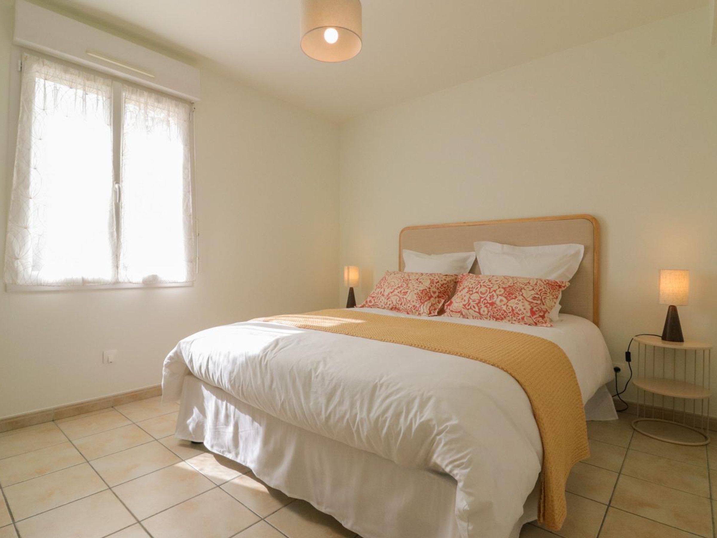 Ein Schlafzimmer mit einem Bett, zwei Nachttischlampen, einem Fenster mit durchsichtigen Vorhängen und einer Leuchte über dem Bett.