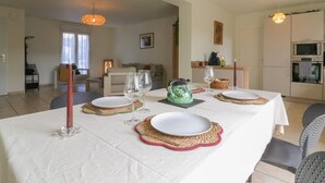 Uma sala de jantar com mesa posta para quatro pessoas, cozinha ao fundo e sala de estar com sofá e abajur.