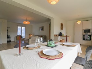 Un comedor con una mesa preparada para cuatro, una cocina al fondo y una sala de estar con un sofá y una lámpara.
