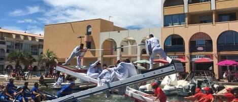 A boat race with participants in white and red uniforms, a waterfront setting with buildings in the background, and spectators watching from the shore.