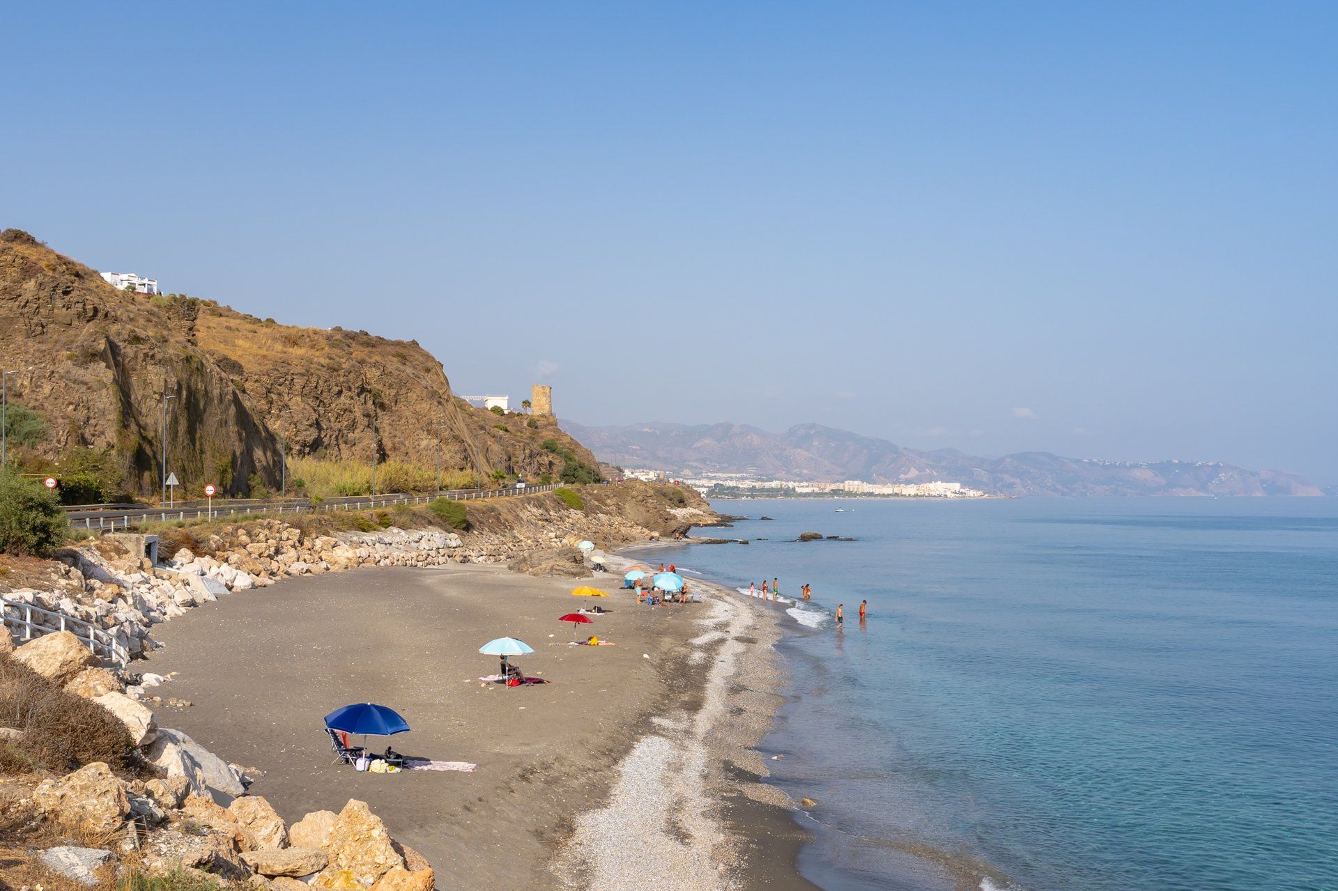 A beach with umbrellas, rocky terrain, and a distant mountain range.