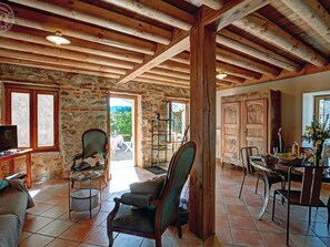 Ein rustikales Interieur mit freiliegenden Holzbalken, Steinwänden, einer Essecke mit Tisch und Stühlen sowie einer gemütlichen Sitzecke mit Sofa und Sesseln.