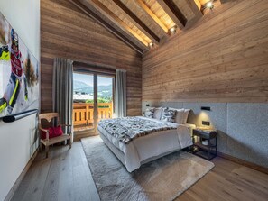 A bedroom with a bed, a nightstand, a chair, a window with a view, and wooden walls. - Apartment in Mont d'Arbois - 6 guests (Megève)