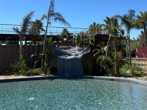 A clear pool with a waterfall feature, surrounded by palm trees and a wooden fence. - Mobile home with AC - 5 guests (Sainte-Marie-la-Mer)