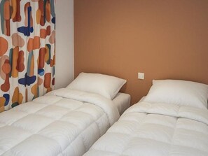 Ein Zimmer mit zwei Einzelbetten, einer schlichten braunen Wand und einem Vorhang mit buntem, abstraktem Muster.