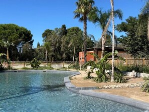 A swimming pool with a curved edge, surrounded by a sandy area with palm trees and a wooden fence. - Air-conditioned mobile home - 6 guests (Sainte-Marie-la-Mer)