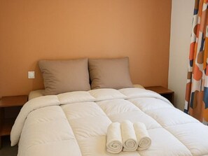 Een opgemaakt bed met een beige dekbed, twee kussens en drie opgerolde handdoeken.