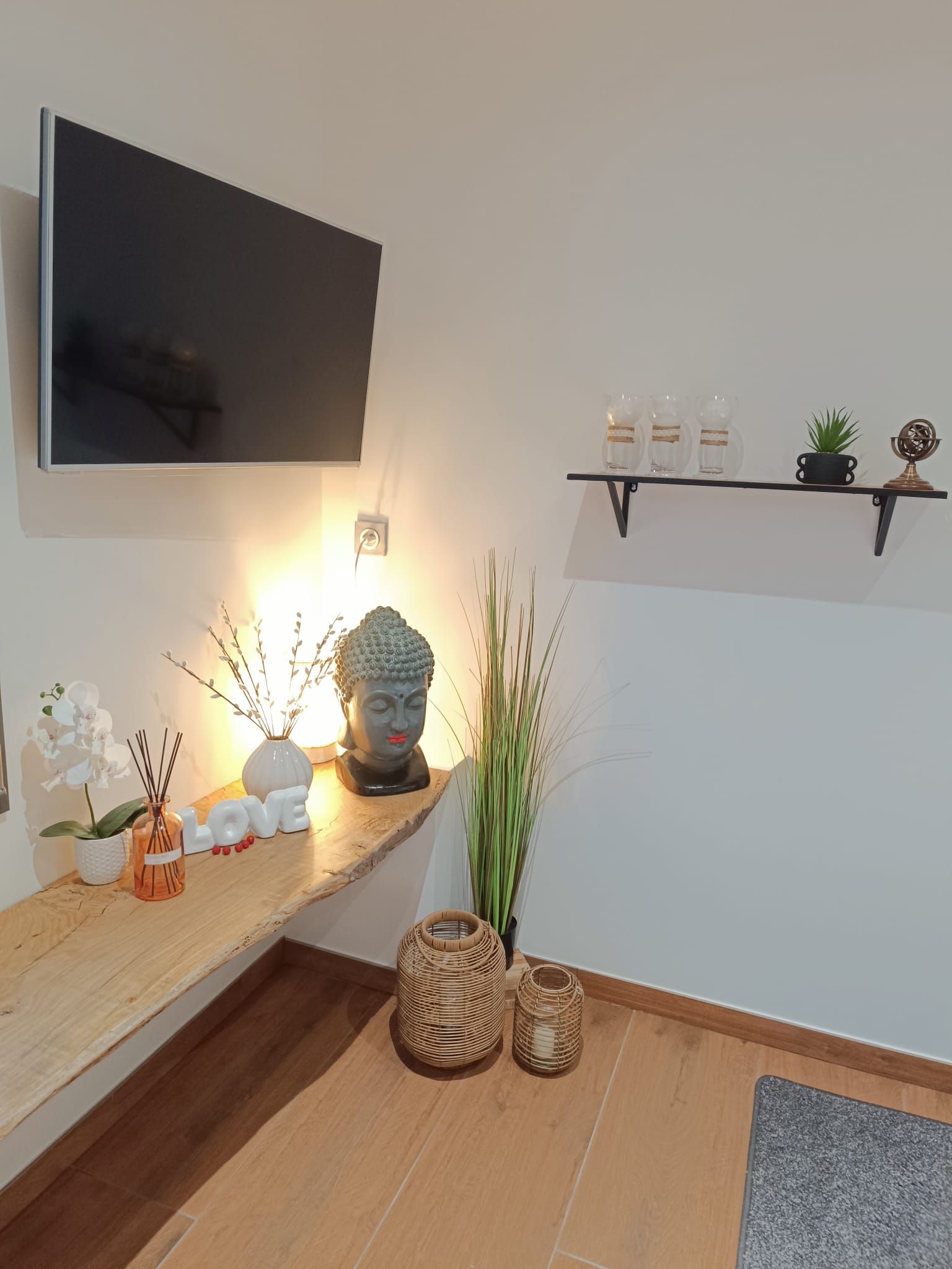 A modern living room with a flat-screen TV mounted on the wall, a wooden shelf with decorative items, a Buddha statue, and a small potted plant.