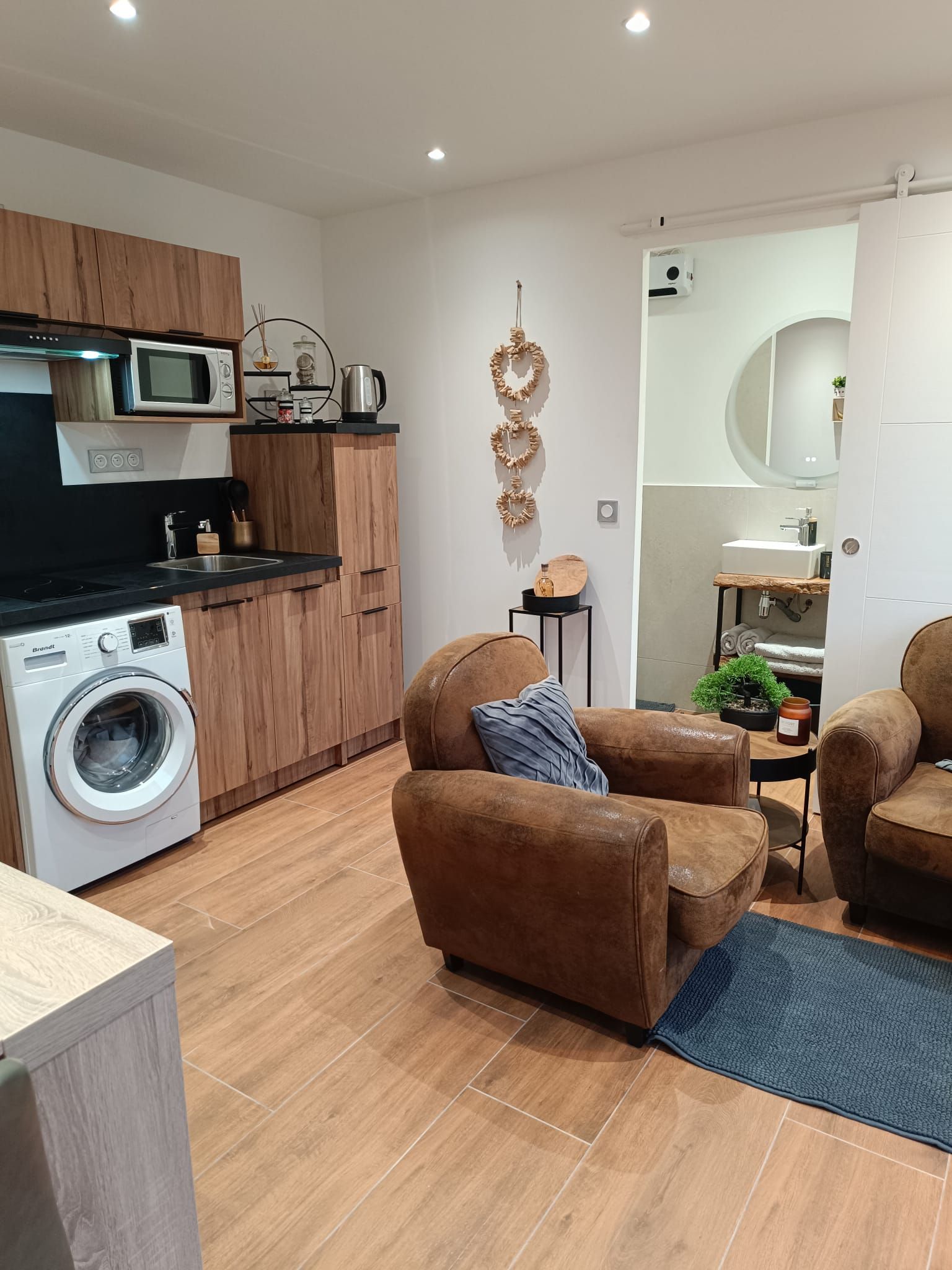 A modern kitchen with a built-in microwave, a washing machine, and a coffee machine. There are two brown sofas and a small table with a plant.