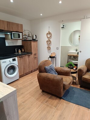 A modern kitchen with a built-in microwave, a washing machine, and a coffee machine. There are two brown sofas and a small table with a plant.