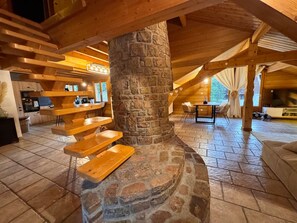 A rustic interior with a stone fireplace, wooden beams, a dining area, and a kitchen with wooden shelves.