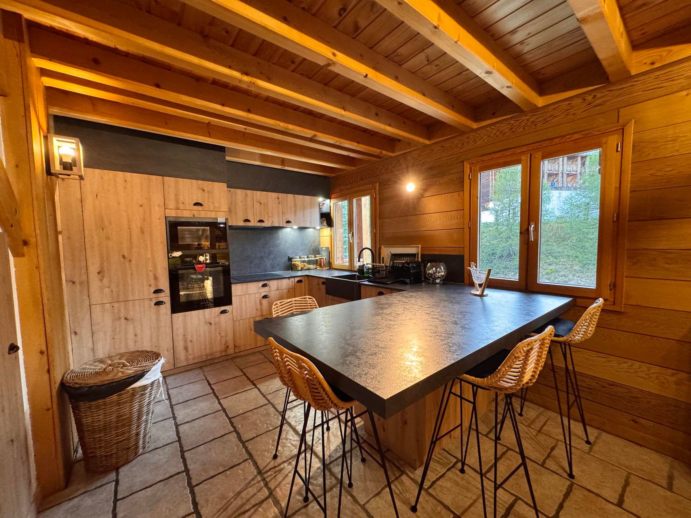 A wooden cabin interior with a kitchen area, a dining table with chairs, and a large window.