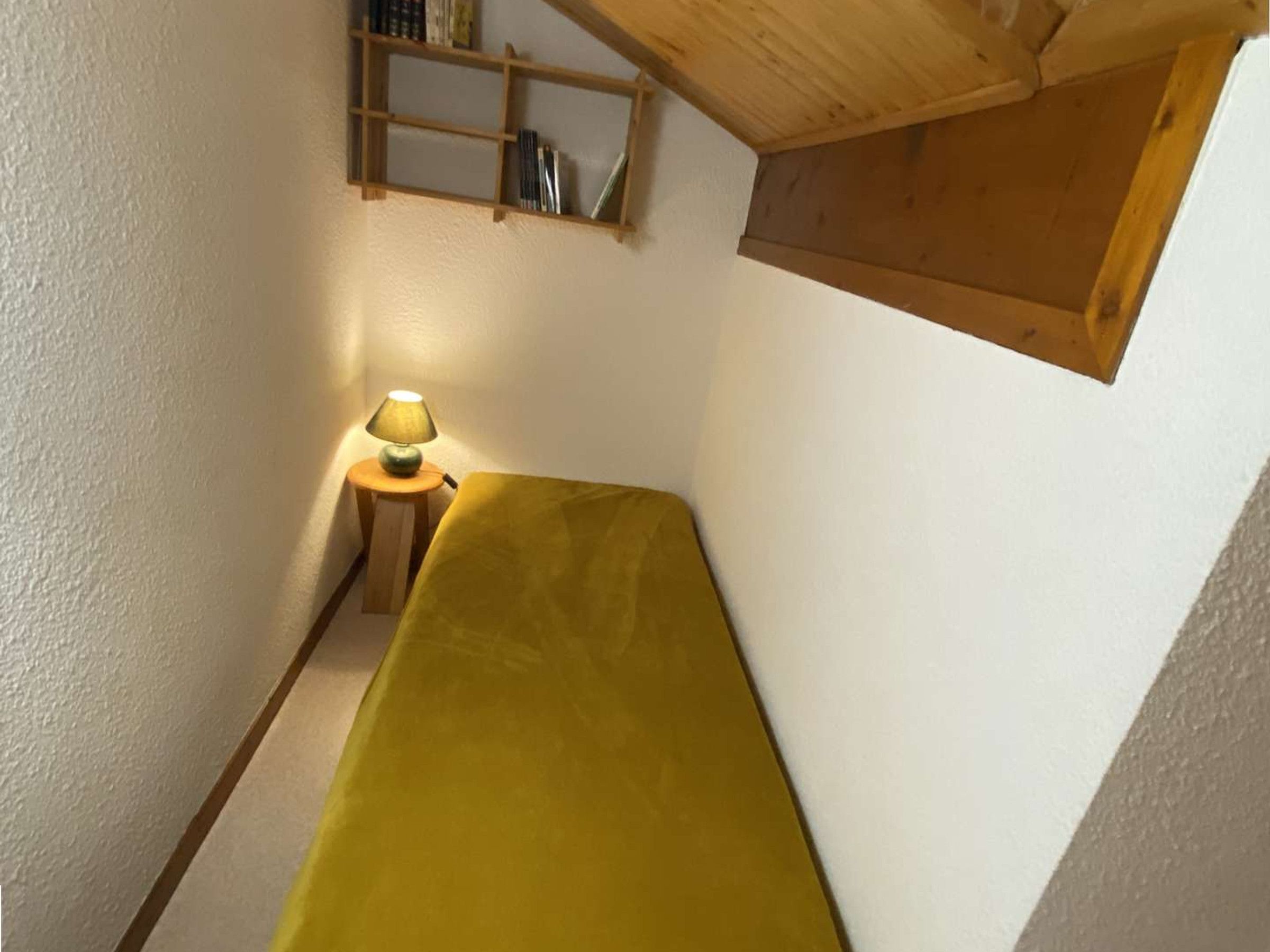 Een kleine kamer met een enkel bed, een nachtkastje met een lamp, en een houten plank boven het bed.