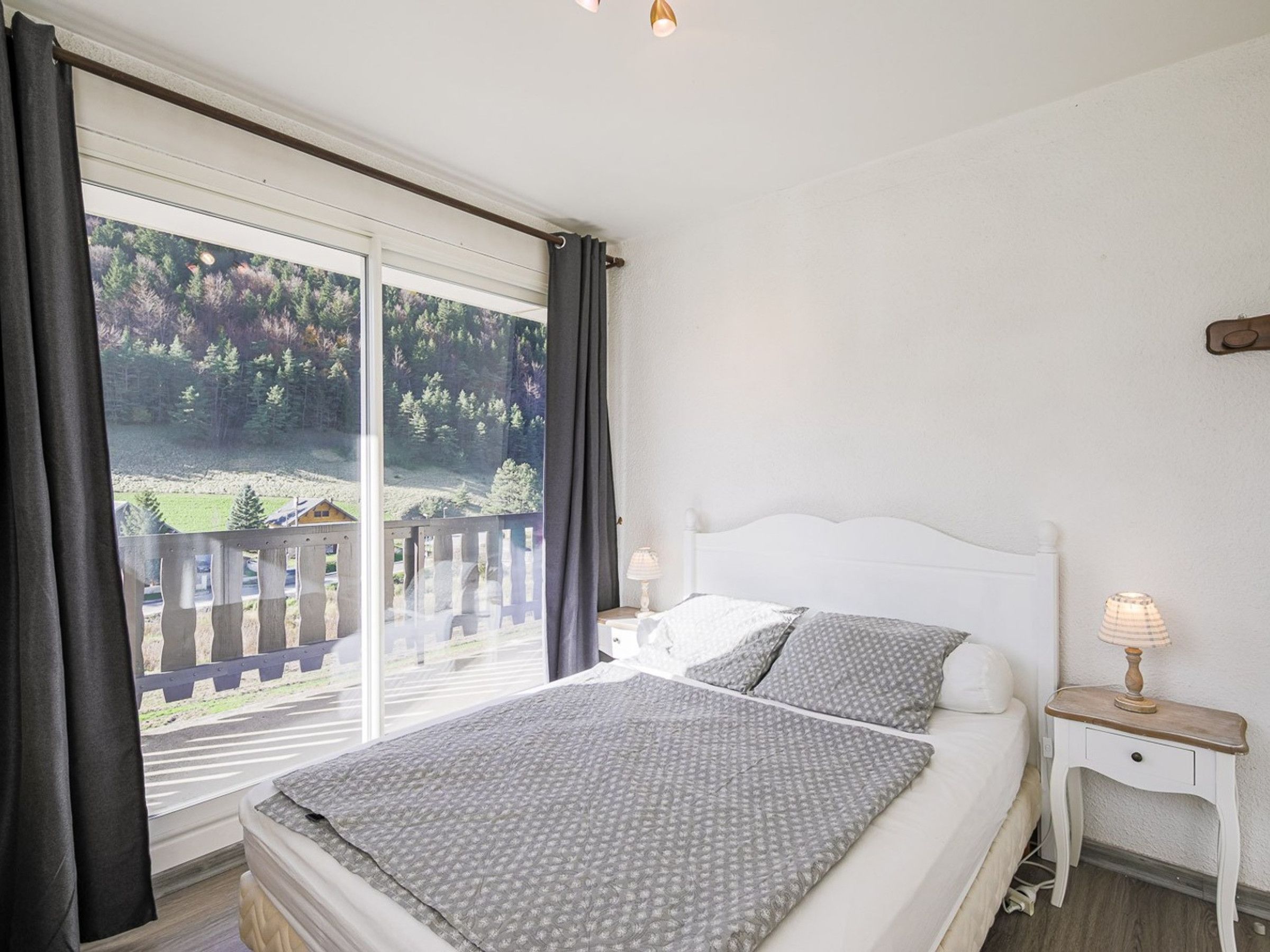 A bedroom with a bed, a nightstand, a lamp, and a view of a forest through a large window.
