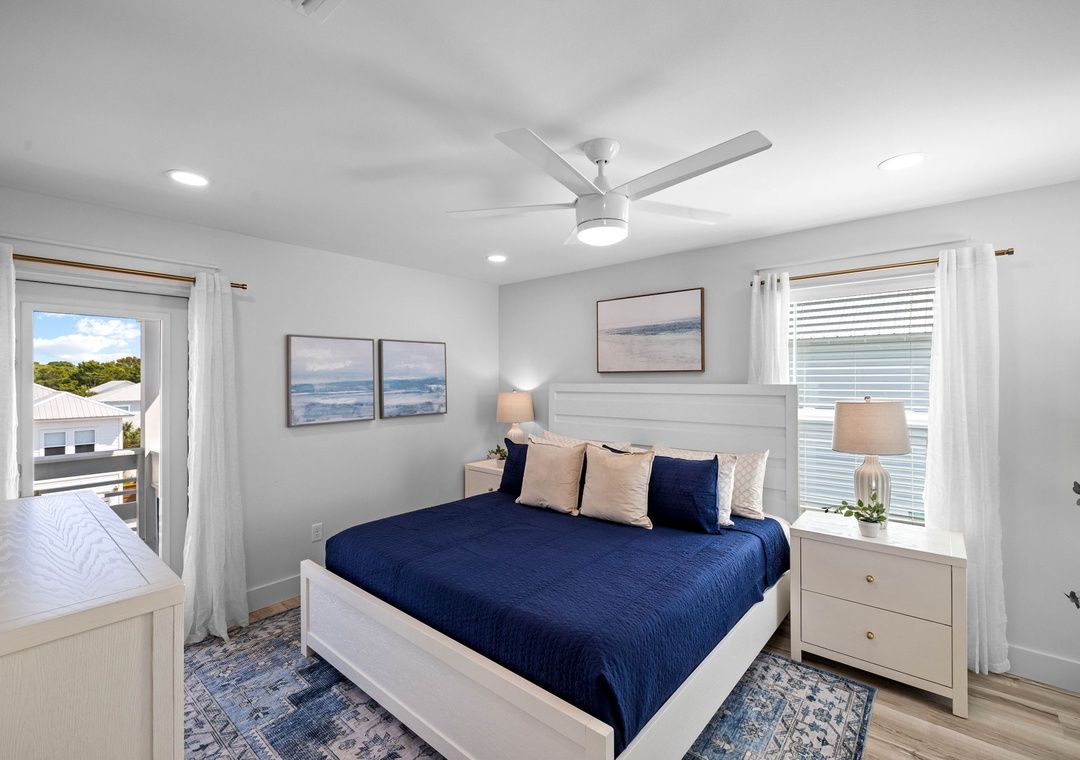 A bedroom with a bed, bedside tables, a ceiling fan, and a view of the outside.