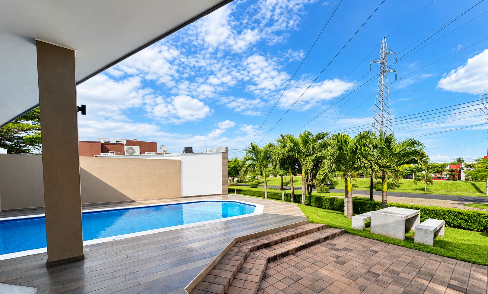 A swimming pool with a tiled deck, a covered area with a white ceiling, and a view of a street with palm trees and a power line tower.